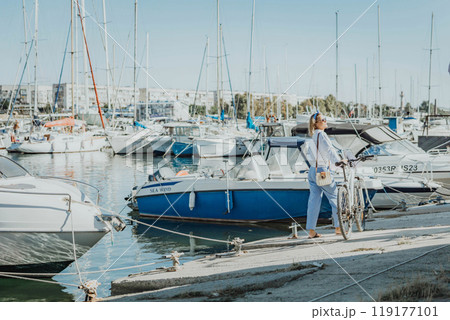 Woman enjoys bike ride along waterfront, Marina surroundings. She is wearing a white shirt and blue jeans, and she has a handbag with her. Capturing outdoor bike ride by waterfront. 119177101