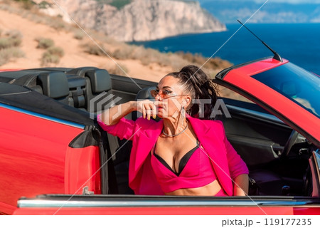A woman in a pink suit is sitting in a red convertible. She is wearing sunglasses and a necklace. A woman in a pink suit is sitting in a red convertible. She is wearing sunglasses and a necklace. 119177235
