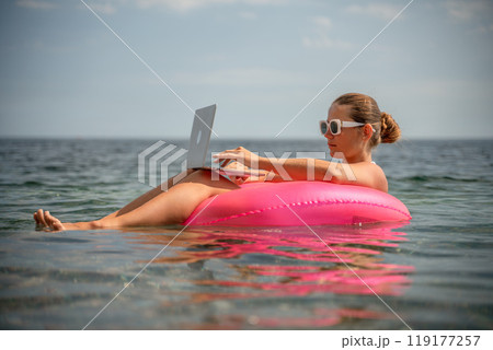 A woman is floating in a pink inflatable raft with a laptop in her hand. She is smiling and she is enjoying her time in the water. 119177257