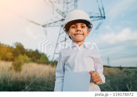 Little kid pretending engineer or manager with safety helmet, with papers documents. Electrical industry profession, serious smart child on transmission power lines background. Little kid pretending engineer or manager with safety helmet, with papers documents. Electrical industry profession, serious smart child on transmission power lines background. 119178072