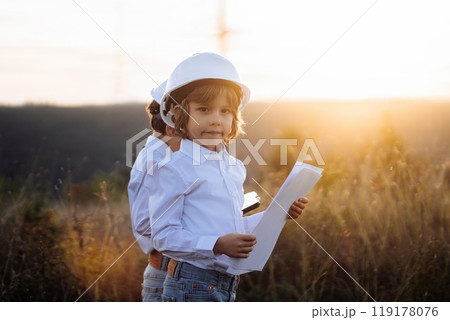 Little boy acting role of engineer or manager with safety helmet. Occupation in electrical industry, serious smart child on electric voltage towers background Little boy acting role of engineer or manager with safety helmet. Occupation in electrical industry, serious smart child on electric voltage towers background 119178076