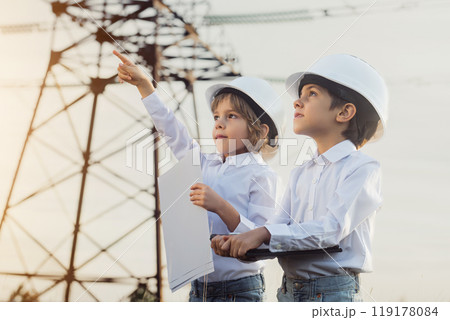 Little kids pretending engineers or managers with safety helmets, reading papers documents. Electrical industry profession, serious smart children on transmission power lines background. Little kids pretending engineers or managers with safety helmets, reading papers documents. Electrical industry profession, serious smart children on transmission power lines background. 119178084