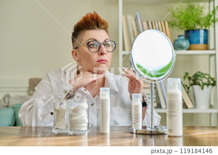 Middle aged woman looking in mirror, using face creams, cosmetic care products Middle aged woman looking in mirror, using face creams, cosmetic care products 119184146