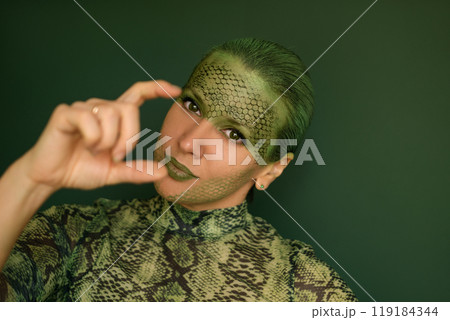 A woman with bright makeup and painted snake scales on her face, gestures with her fingers. A character in the guise of a reptile, a symbol of the 2025 new year on a green studio background A woman with bright makeup and painted snake scales on her face, gestures with her fingers. A character in the guise of a reptile, a symbol of the 2025 new year on a green studio background 119184344
