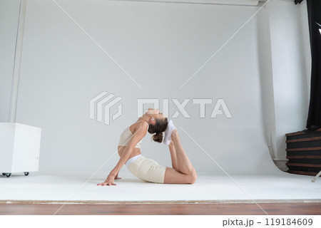 Dressed in a beige sports outfit, a woman bends backward in a graceful yoga pose against a plain white backdrop. This image captures the essence of yoga practice and the pursuit of a balanced Dressed in a beige sports outfit, a woman bends backward in a graceful yoga pose against a plain white backdrop. This image captures the essence of yoga practice and the pursuit of a balanced 119184609