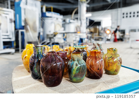 Many colorful glass vases at glass blowing manufacture factory  with furnace equipment set on background. Handmade handcrafted blowing glassware design production workshop Europe 119185357