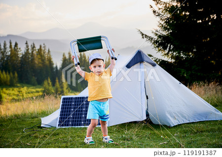 Young child playing near tourist tent with photovoltaic solar panel in summer. On background of mountains and trees under clear blue sky. Integration of renewable energy in outdoor camping activities. Young child playing near tourist tent with photovoltaic solar panel in summer. On background of mountains and trees under clear blue sky. Integration of renewable energy in outdoor camping activities. 119191387