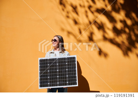Woman holding solar panel for charging, stands against vibrant orange wall. Smiling girl wears sunglasses, white top. Artistic shadows cast by nearby trees branches on wall. 119191391