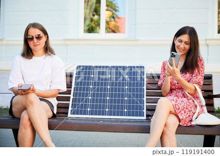 Young girl can't charge her phone because her friend used all energy. Confused girl doesn't know how to charge phone from solar battery. Two female sitting on bench with solar panel between them. Young girl can't charge her phone because her friend used all energy. Confused girl doesn't know how to charge phone from solar battery. Two female sitting on bench with solar panel between them. 119191400