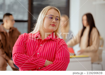 Woman in a pink striped shirt stands in front of a window with her arms crossed, people in the team work in the background Woman in a pink striped shirt stands in front of a window with her arms crossed, people in the team work in the background 119200639