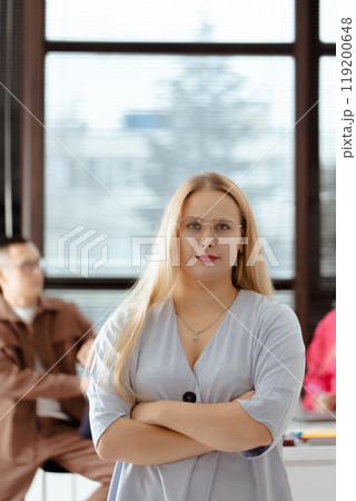 A serious woman with blonde hair and glasses stands in front of a window with her arms crossed, people in the team work in the background A serious woman with blonde hair and glasses stands in front of a window with her arms crossed, people in the team work in the background 119200648