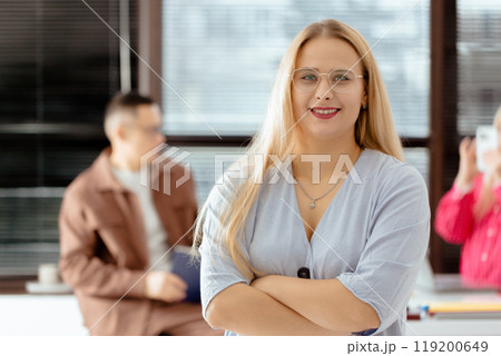 A happy successful woman with blonde hair and glasses stands in front of a window with her arms crossed A happy successful woman with blonde hair and glasses stands in front of a window with her arms crossed 119200649