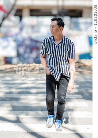 A stylish young man confidently walking in an urban setting wearing a striped shirt 119201038