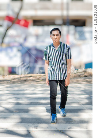 A confident young man is walking in an urban setting while wearing a striped shirt 119201039