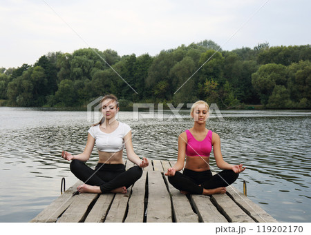 Two teenage girls doing yoga exercises for concentration and relaxation on a wooden pier 119202170