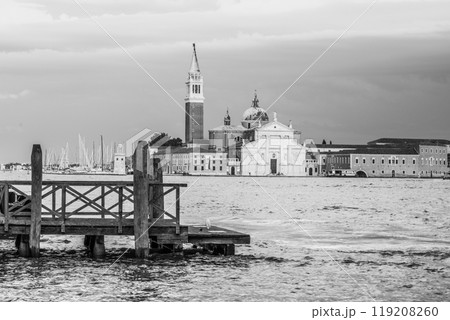 The iconic San Giorgio Maggiore stands majestically against a cloudy sky, viewed from the shores of Venice. Boats gently bob in the water, adding to the serene atmosphere of this historic location. 119208260