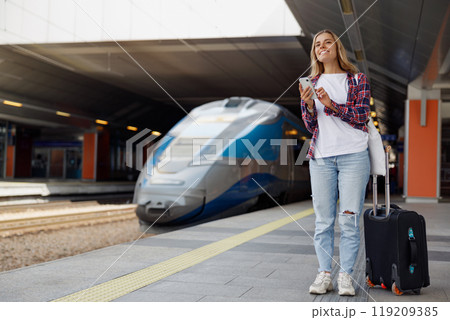 A Young Woman Standing at a Busy Train Station with Her Luggage and Smartphone in Hand 119209385
