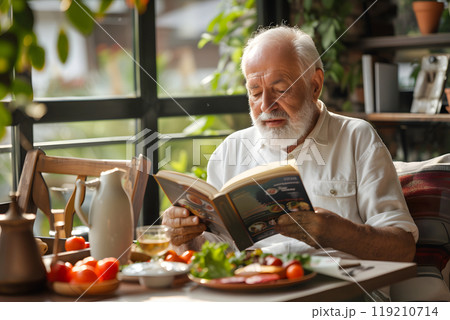 An old man reads a book at a table with his meal 119210714