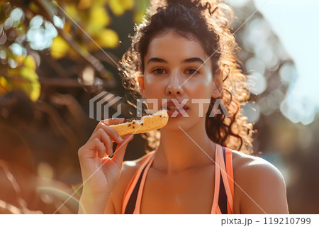 A woman in a sports bra eats a snack while outdoors 119210799