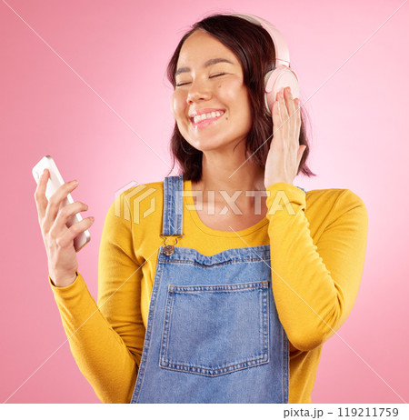 Woman, headphones and cellphone for smile, studio or thinking for sound, idea and relax by pink background. Happy gen z girl, Japanese student and music app for listening with streaming subscription Woman, headphones and cellphone for smile, studio or thinking for sound, idea and relax by pink background. Happy gen z girl, Japanese student and music app for listening with streaming subscription 119211759
