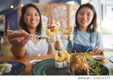 Chopsticks, girl friends and shrimp sushi at a table with salmon and Japanese cuisine food at restaurant. Young women, eating and tempura prawn with fish for lunch and meal on a plate with a smile Chopsticks, girl friends and shrimp sushi at a table with salmon and Japanese cuisine food at restaurant. Young women, eating and tempura prawn with fish for lunch and meal on a plate with a smile 119212577