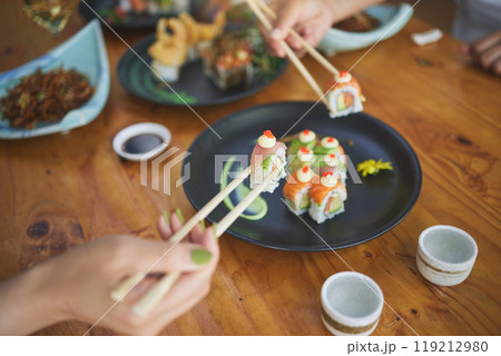 Sushi, hands and eating food with chopsticks at restaurant for nutrition at table. Closeup of people with wooden sticks for dining, Japanese culture and cuisine while sharing with creativity on plate 119212980