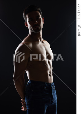 Bodybuilder man, shirtless and muscle in studio for wellness, healthy body and black background. Young guy, fitness and silhouette in jeans, portrait and strong for fashion, health or topless for art Bodybuilder man, shirtless and muscle in studio for wellness, healthy body and black background. Young guy, fitness and silhouette in jeans, portrait and strong for fashion, health or topless for art 119215827