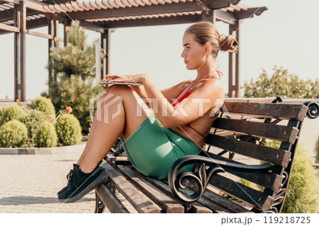 Woman Workout Bench Park - A woman in workout clothes sits on a park bench during the day. She is taking a break from exercise. Woman Workout Bench Park - A woman in workout clothes sits on a park bench during the day. She is taking a break from exercise. 119218725