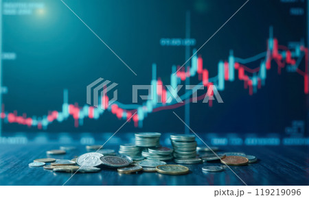 Stacked coins in focus with scattered currency on table against blurred stock market chart background showing investment growth and financial trading analysis 119219096