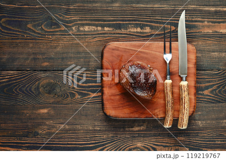 A perfectly cooked steak rests on a wooden cutting board with utensils nearby 119219767