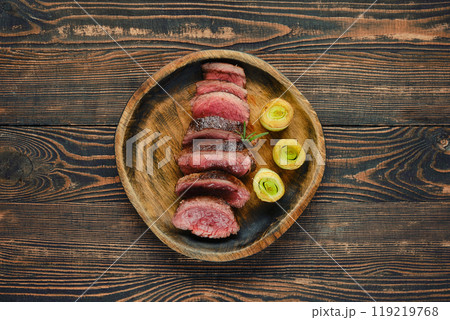 Sliced roast beef steak with roasted leek served on a wooden plate against a rustic background Sliced roast beef steak with roasted leek served on a wooden plate against a rustic background 119219768