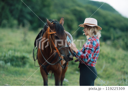Happy blonde with horse in forest. Woman and a horse walking through the field during the day. Dressed in a plaid shirt and black leggings. 119225309