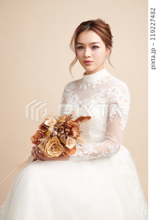 Beautiful young asian woman in wedding dress holding bouquet on beige background, 119227482
