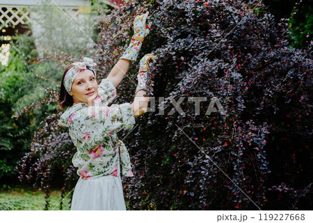 Woman dressed in a floral gardening outfit is pruning a large shrub in a lush garden Woman dressed in a floral gardening outfit is pruning a large shrub in a lush garden 119227668