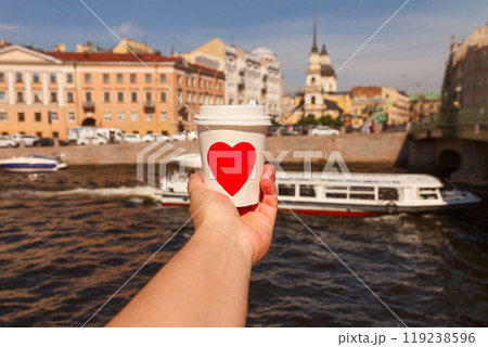 white cup of drink with red heart in woman's palm near embankment of Fontanka river with ship in St. Petersburg 119238596