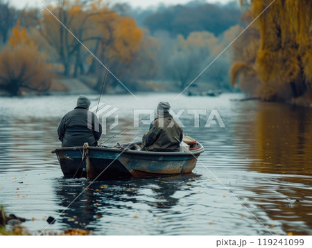 Two individuals sit in a small boat, fishing calmly on a river surrounded by vibrant autumn trees and reflecting water under an overcast sky. 119241099