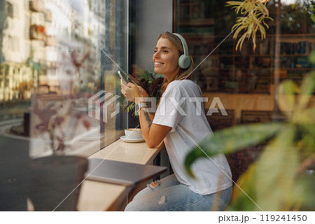A Woman Happily Enjoying Her Coffee While Listening to Music Through Headphones in a Modern Cafe 119241450