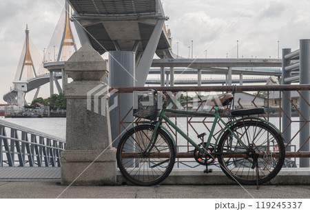 An old bicycle parked on the side in the ferry pier with Bhumibol suspension bridge cross over Chao Phraya River in the background. An old bicycle parked on the side in the ferry pier with Bhumibol suspension bridge cross over Chao Phraya River in the background. 119245337