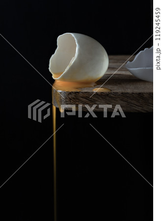 Conceptual image of a broken white eggshell with dripping egg yolk on black background. Conceptual image of a broken white eggshell with dripping egg yolk on black background. 119245459