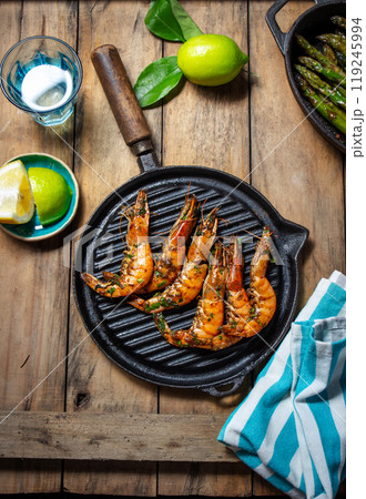 Grilled shrimp on a black grill pan on a rustic wooden table, with limes, a blue-striped cloth Grilled shrimp on a black grill pan on a rustic wooden table, with limes, a blue-striped cloth 119245994