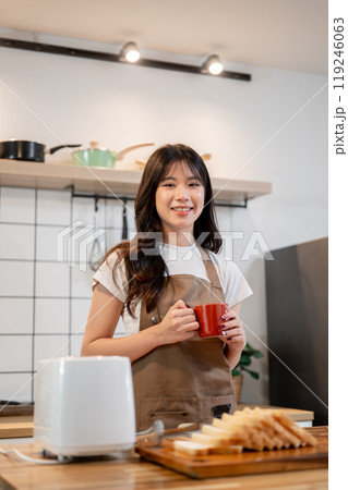 A beautiful Asian woman preparing her breakfast in the kitchen, making toast and coffee. 119246063