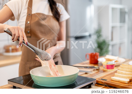 A woman holding tongs, flipping a slice of bacon in a pan while making her breakfast in the kitchen. A woman holding tongs, flipping a slice of bacon in a pan while making her breakfast in the kitchen. 119246076