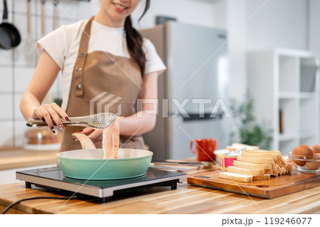 A woman holding tongs, flipping a slice of bacon in a pan while making her breakfast in the kitchen. A woman holding tongs, flipping a slice of bacon in a pan while making her breakfast in the kitchen. 119246077
