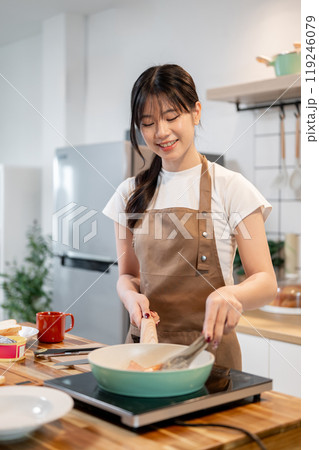 A beautiful, happy Asian woman frying bacons in a pan while making her breakfast in the kitchen. A beautiful, happy Asian woman frying bacons in a pan while making her breakfast in the kitchen. 119246079