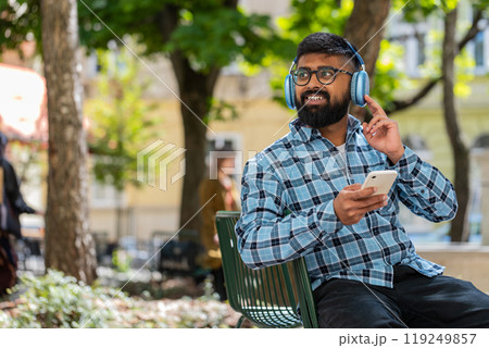 Happy Indian man dancing in wireless headphones listening favorite energetic music on city street 119249857