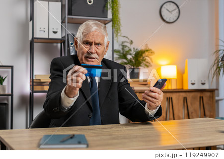 Caucasian angry senior businessman trying to pay online with smartphone blocked credit card at home Caucasian angry senior businessman trying to pay online with smartphone blocked credit card at home 119249907