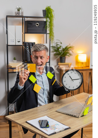 Frustrated young businessman in formal suit with clock and sticky notes sitting at home office desk Frustrated young businessman in formal suit with clock and sticky notes sitting at home office desk 119249935