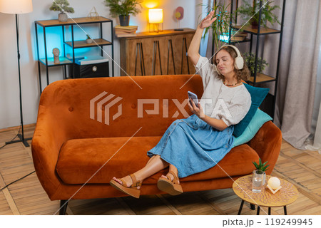 Happy overjoyed young woman in wireless headphones using smartphone relaxing sitting on sofa at home 119249945