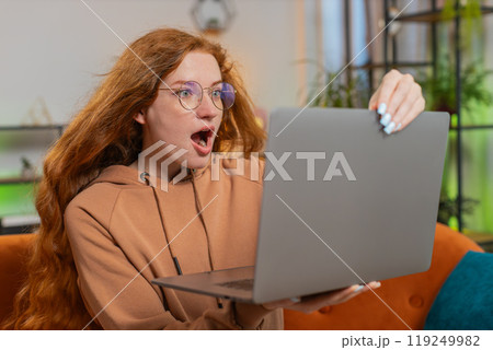 Surprised Caucasian redhead woman with windswept hair using laptop sitting on couch at home 119249982