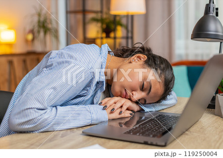 Bored tired sleepy young Hindu woman working on laptop netbook and sleeps at home office desk 119250004